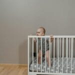 Niemowlę w szarym pajacyku stojące w białym łóżeczku w pokoju