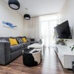 Szara sofa z żółtymi poduszkami w jasnym salonie z telewizorem na ścianie